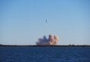 A rocket launching into a clear blue sky over the ocean, with smoke rising from the launch site