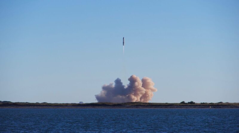 A rocket launching into a clear blue sky over the ocean, with smoke rising from the launch site