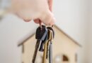 A hand holding house keys in front of a miniature wooden house